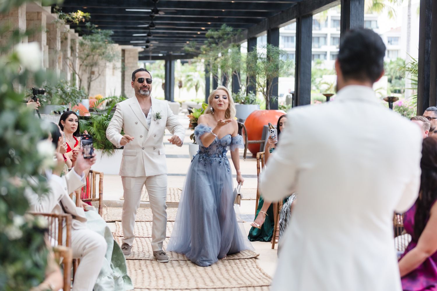 Couple walks down aisle at outdoor wedding ceremony, bride in blue gown, groom in white suit, lush greenery backdrop.