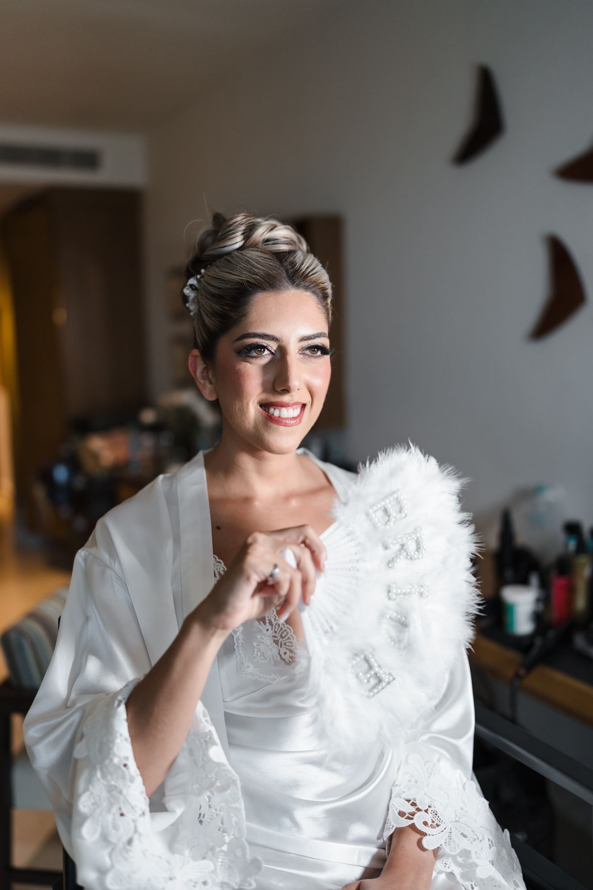 Smiling bride in white satin robe holding a feathered fan while getting ready indoors.