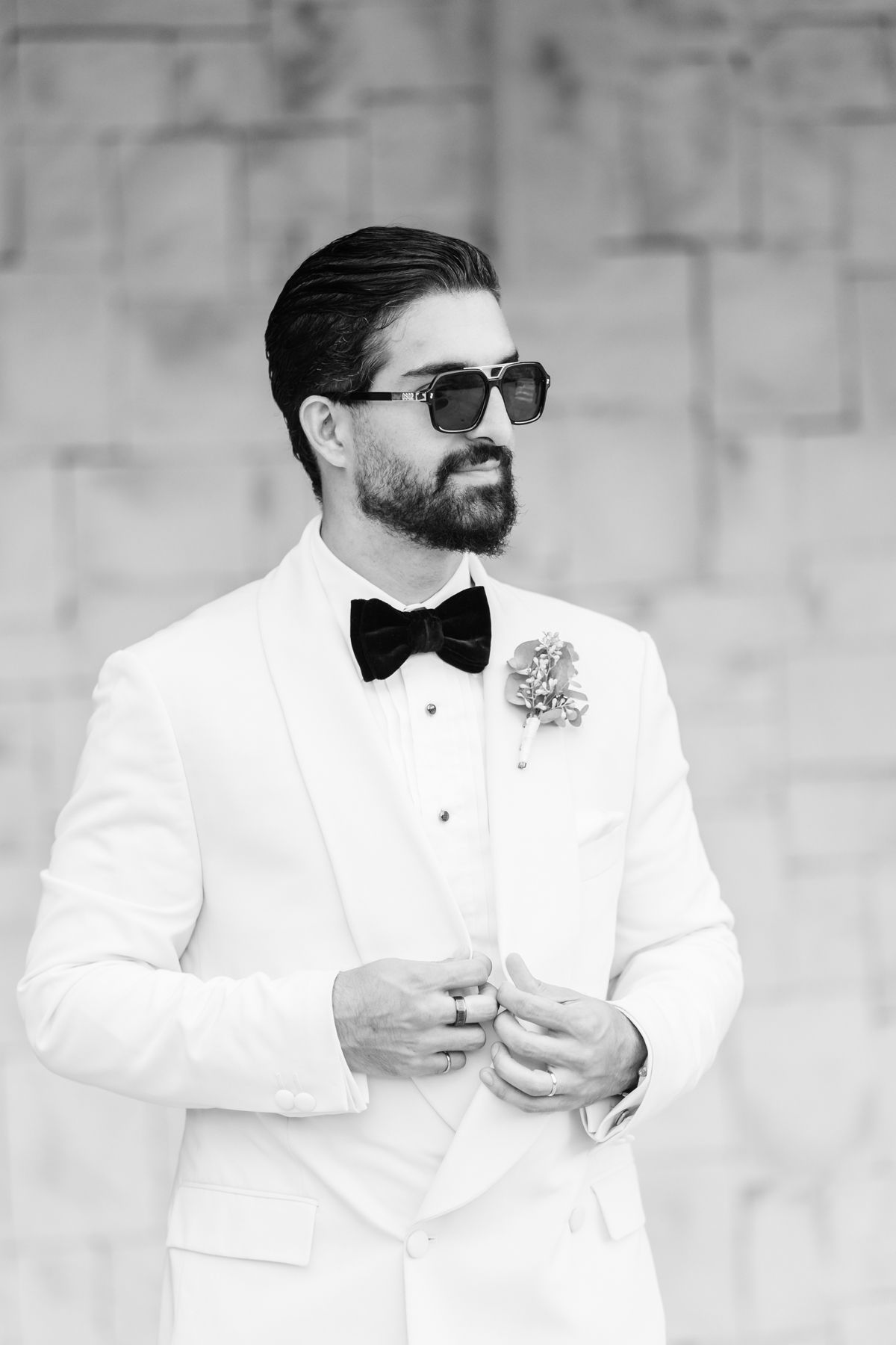 Dapper groom in white tuxedo and black bow tie with boutonniere, wearing sunglasses outdoors.
