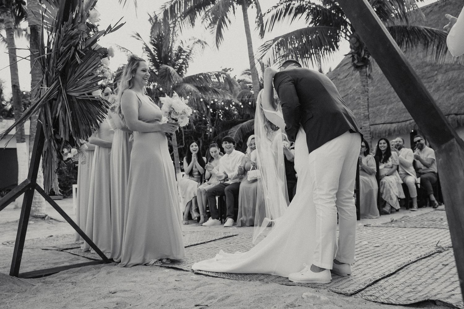 Groom bows before bride during outdoor beach wedding ceremony with guests seated among palm trees.