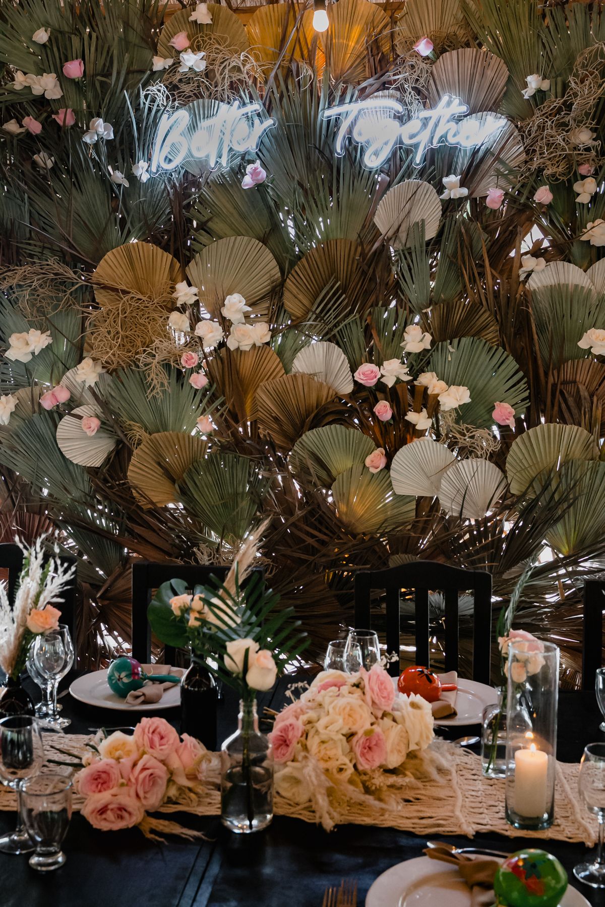 Elegant wedding reception table with pink roses, candles, dried palm fans, and a glowing neon sign backdrop.