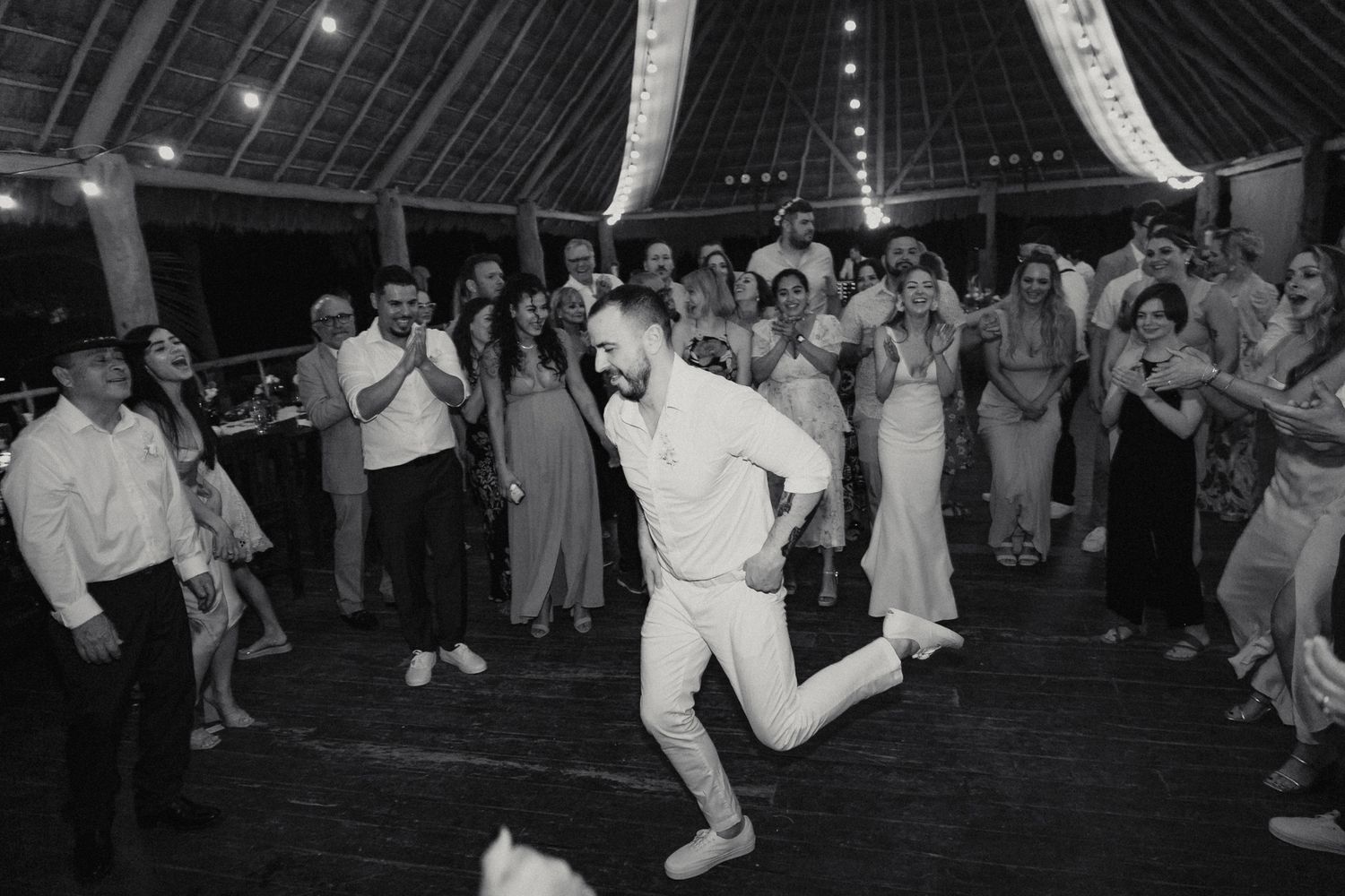 Groom dances energetically for cheering wedding guests on reception dance floor in black and white.
