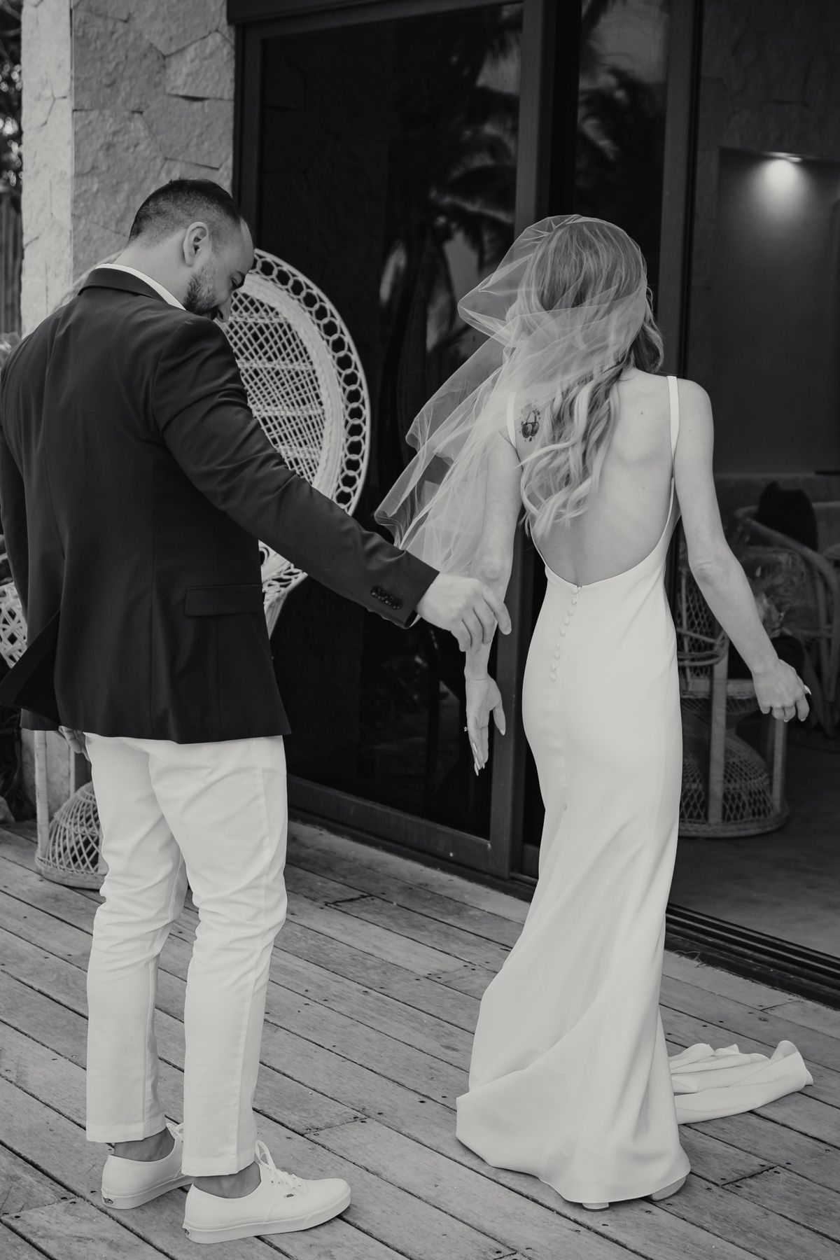 Black and white photo of groom guiding bride in backless wedding gown on wooden deck.