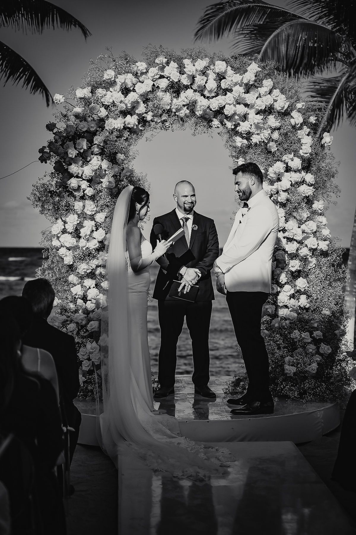 Black and white photo of a couple exchanging vows under a floral arch at a beach wedding ceremony.