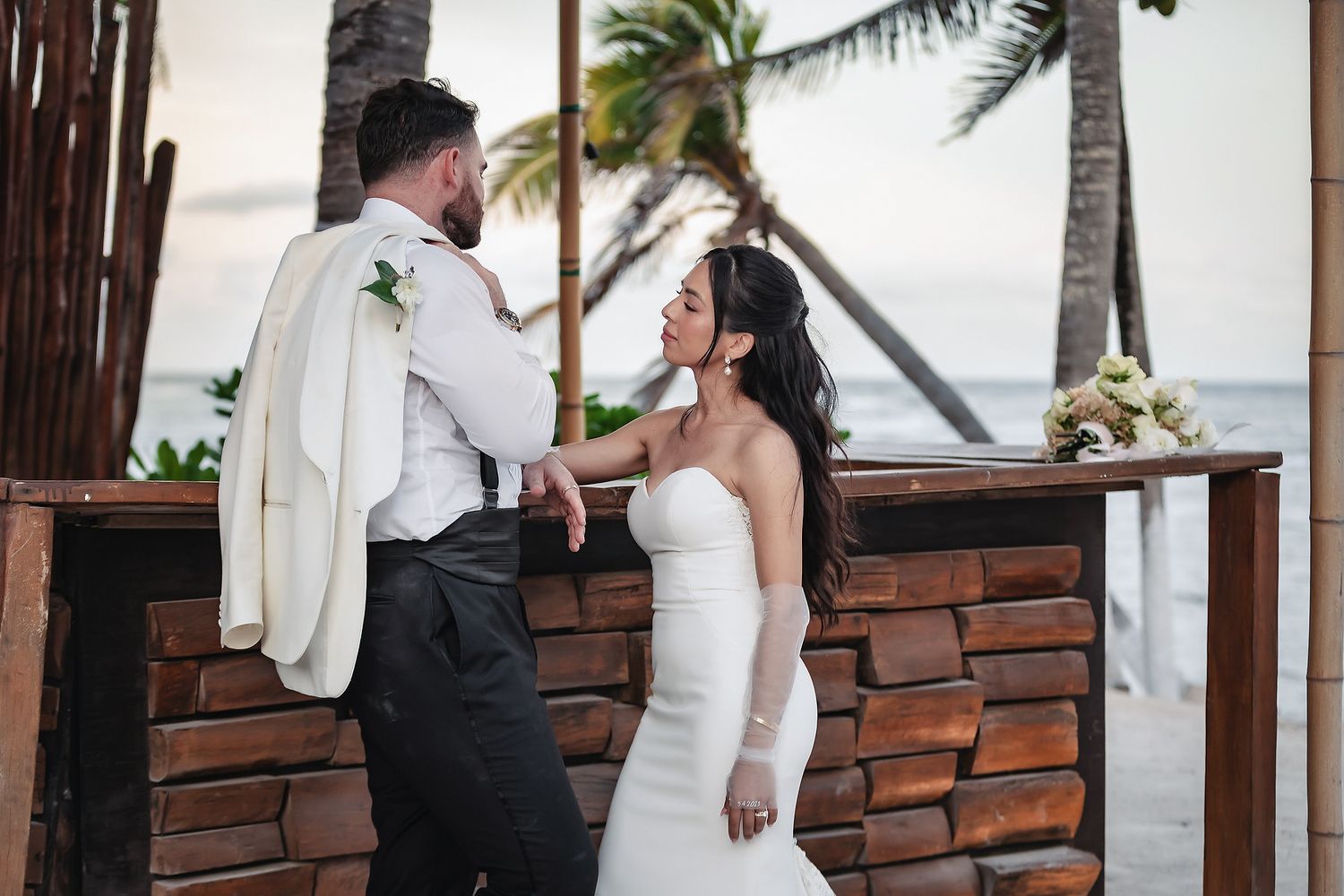 Bride and groom share romantic moment at beachside stone altar surrounded by palm trees at dusk.