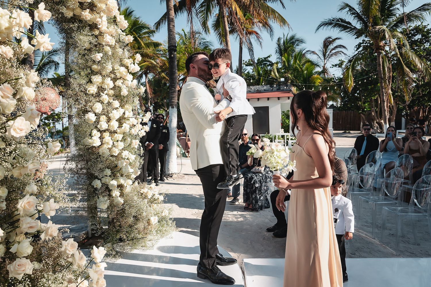 Elegant outdoor wedding ceremony with groom holding child, floral arch, and bridesmaid in blush gown, tropical setting.