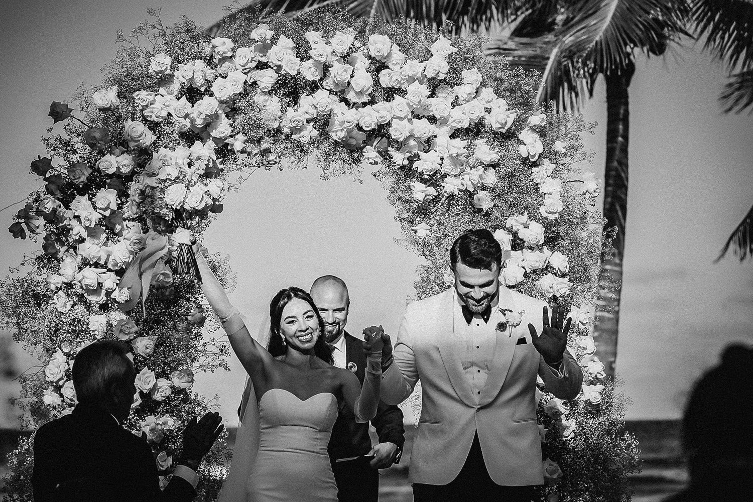 Black and white photo of joyful newlyweds celebrating under floral arch at outdoor wedding ceremony.