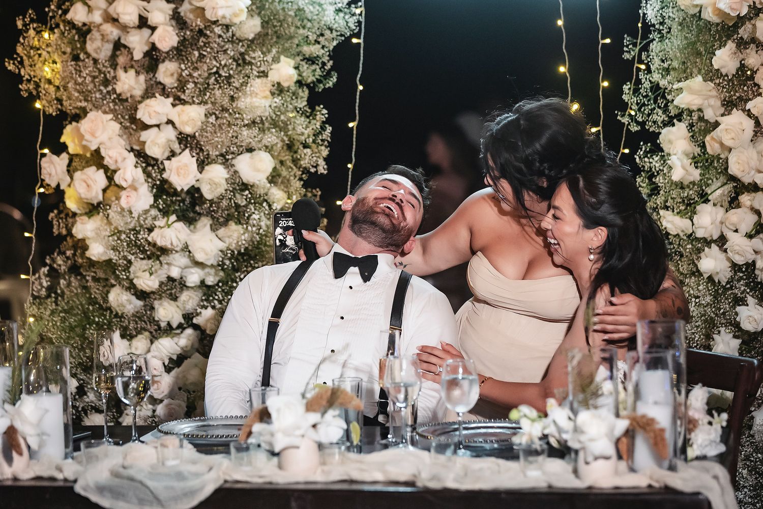 Joyful groom laughing with bride and guest at floral-decorated sweetheart table during wedding reception.