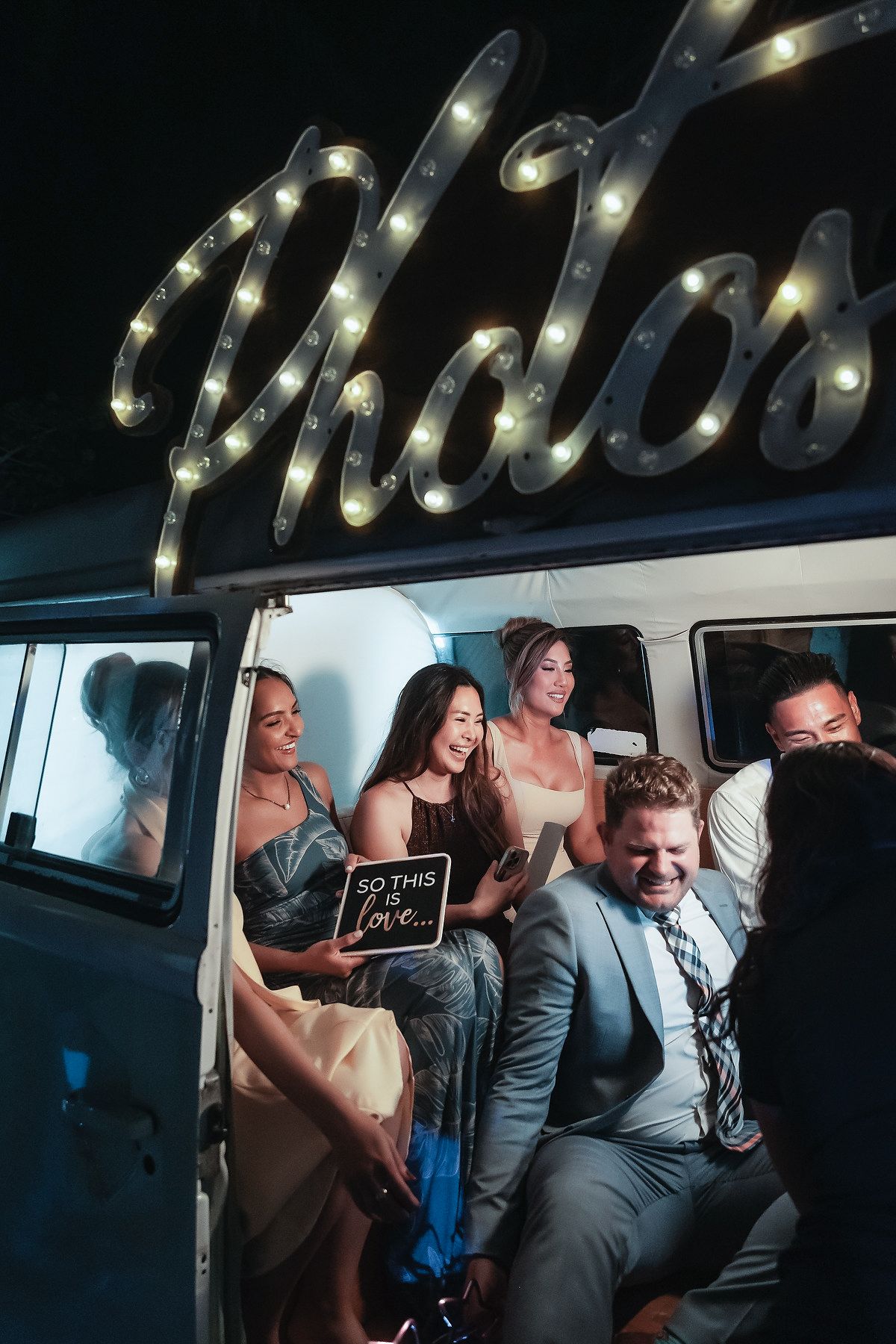 Wedding guests laugh and pose inside vintage photo booth van decorated with marquee lights at reception.