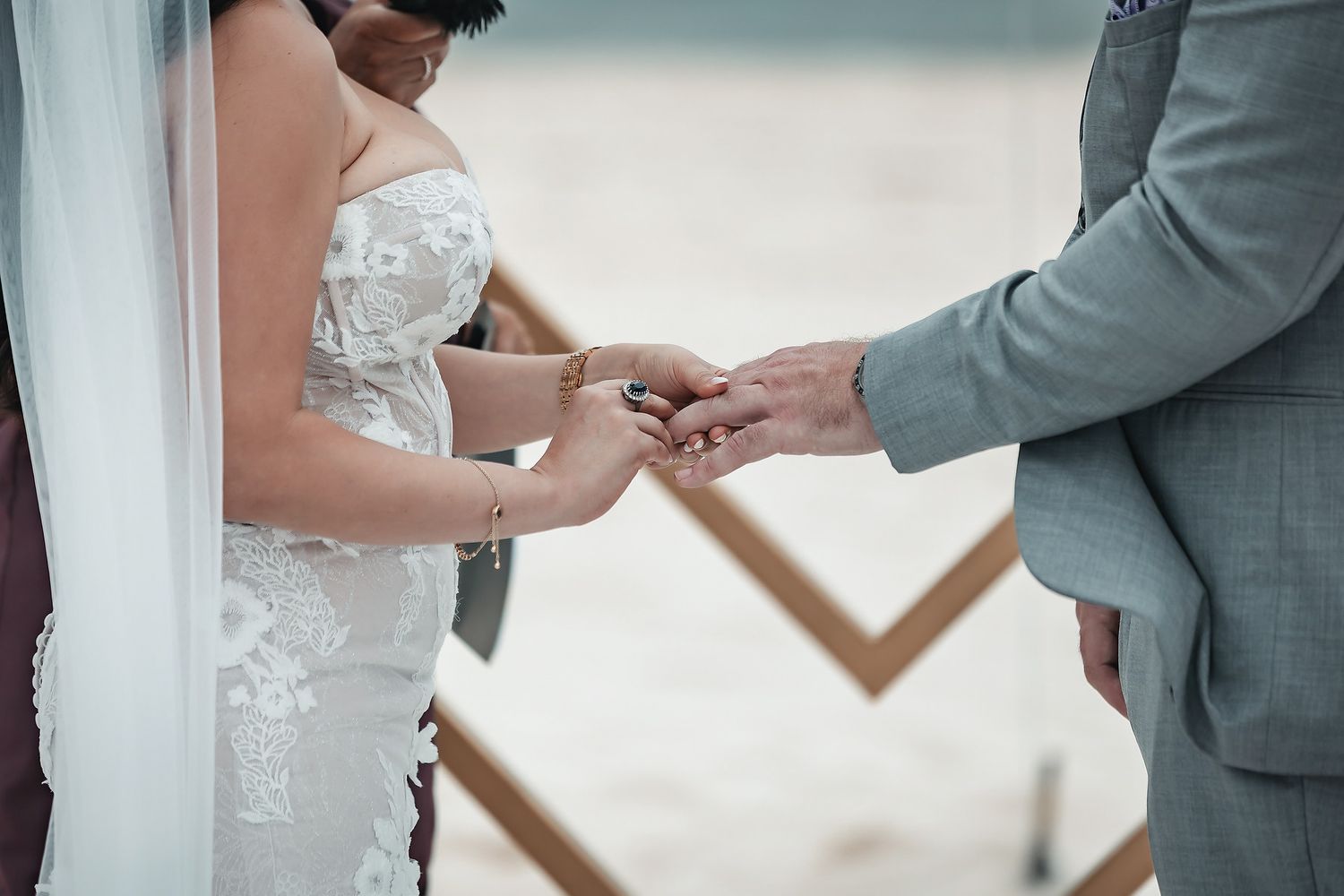 Bride and groom exchanging wedding rings during a beach ceremony, wearing a lace gown and gray suit.
