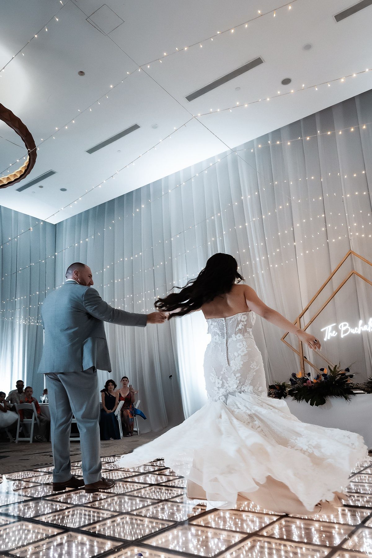 Bride and groom share their first dance on a sparkling dance floor surrounded by fairy lights at an elegant wedding reception.