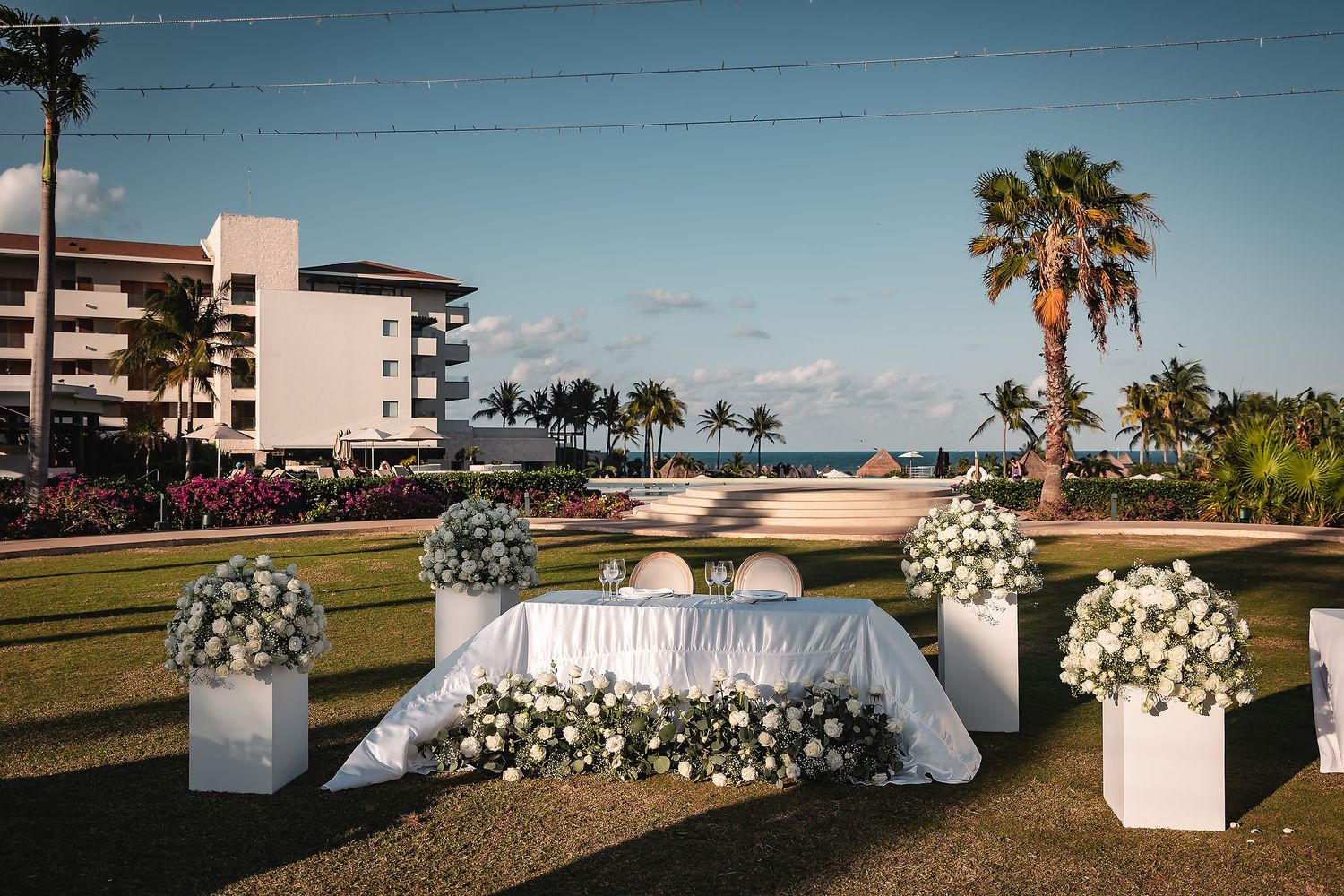 Elegant outdoor wedding reception setup with white floral arrangements, draped sweetheart table, palm trees, and ocean view.