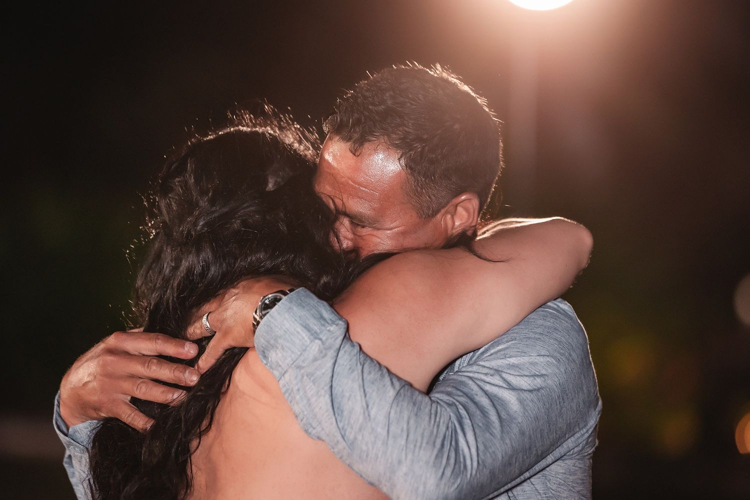 A couple shares a warm embrace under a glowing light at night, capturing a tender romantic moment.