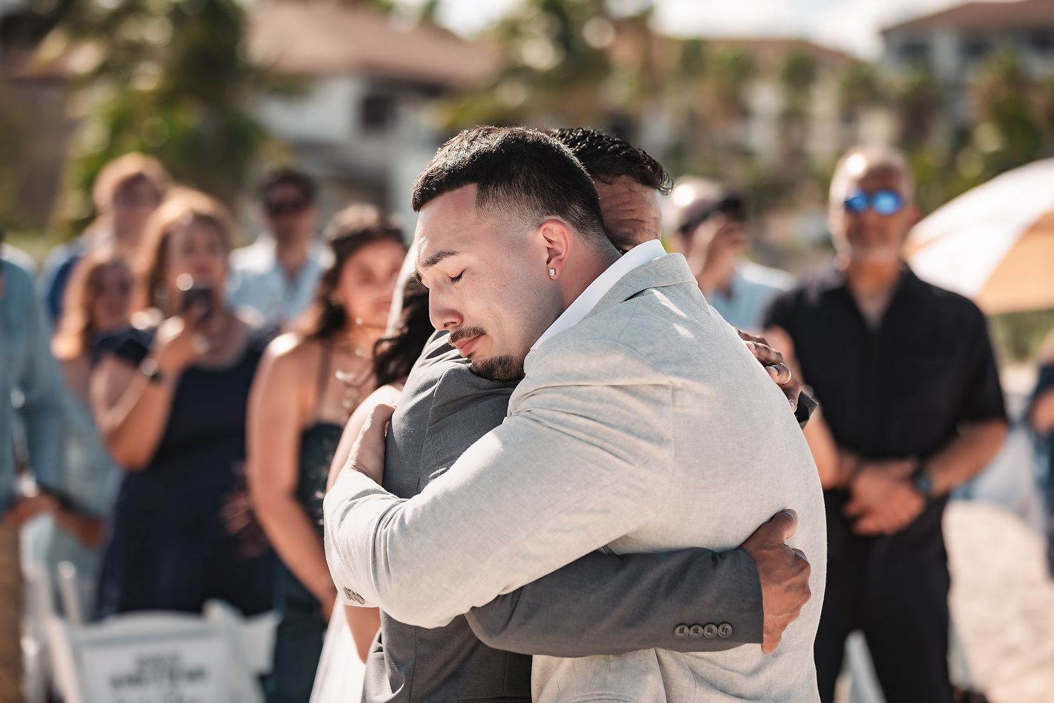 Two men share an emotional hug at an outdoor beach wedding ceremony with guests watching in background.