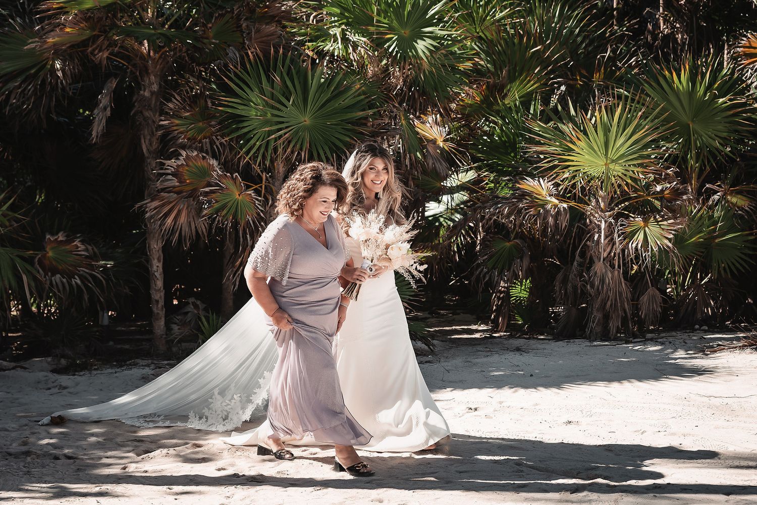 Bride and companion laugh together on sandy beach surrounded by lush tropical palm trees.