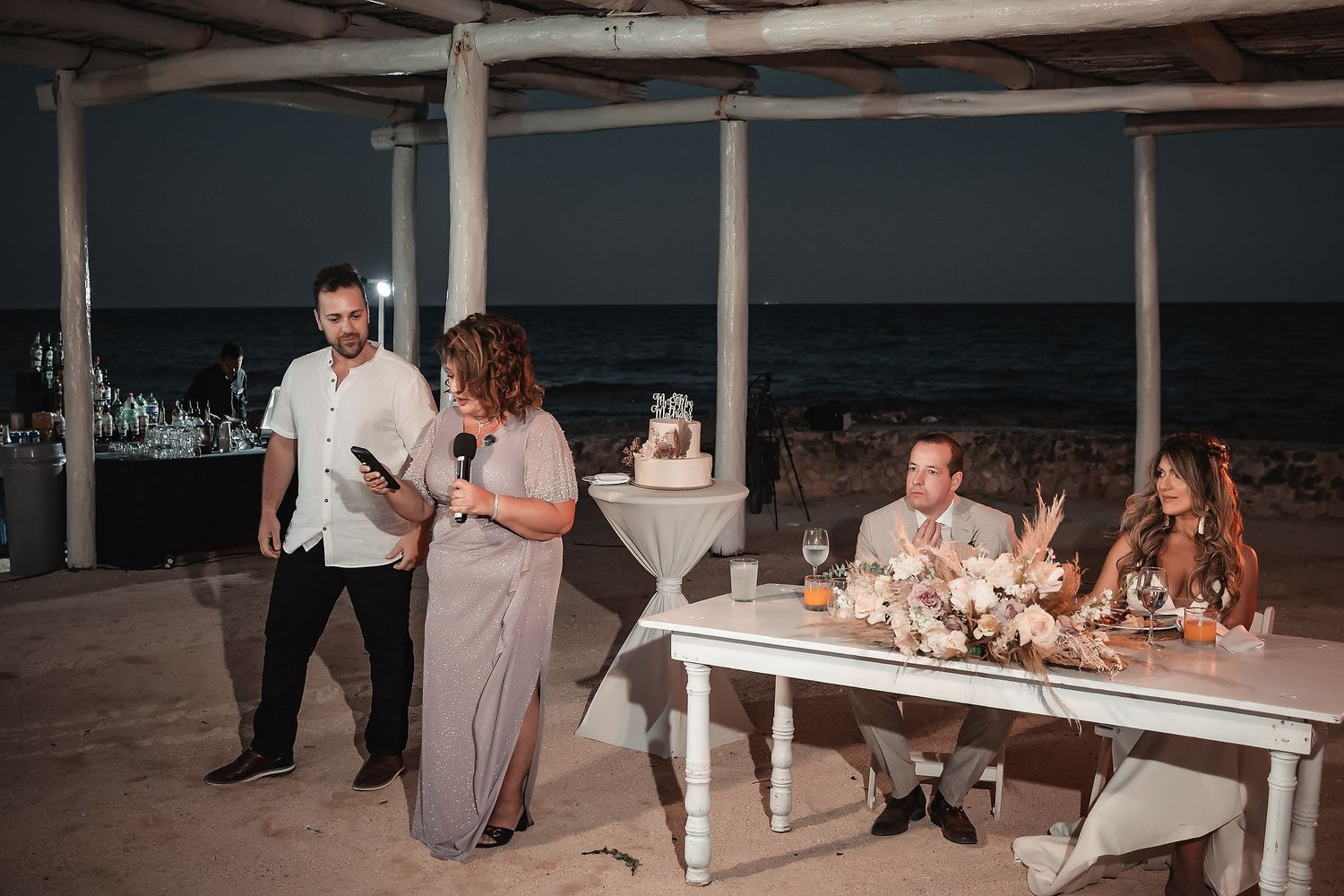 Couple giving speech at outdoor beach wedding reception at night with ocean backdrop.