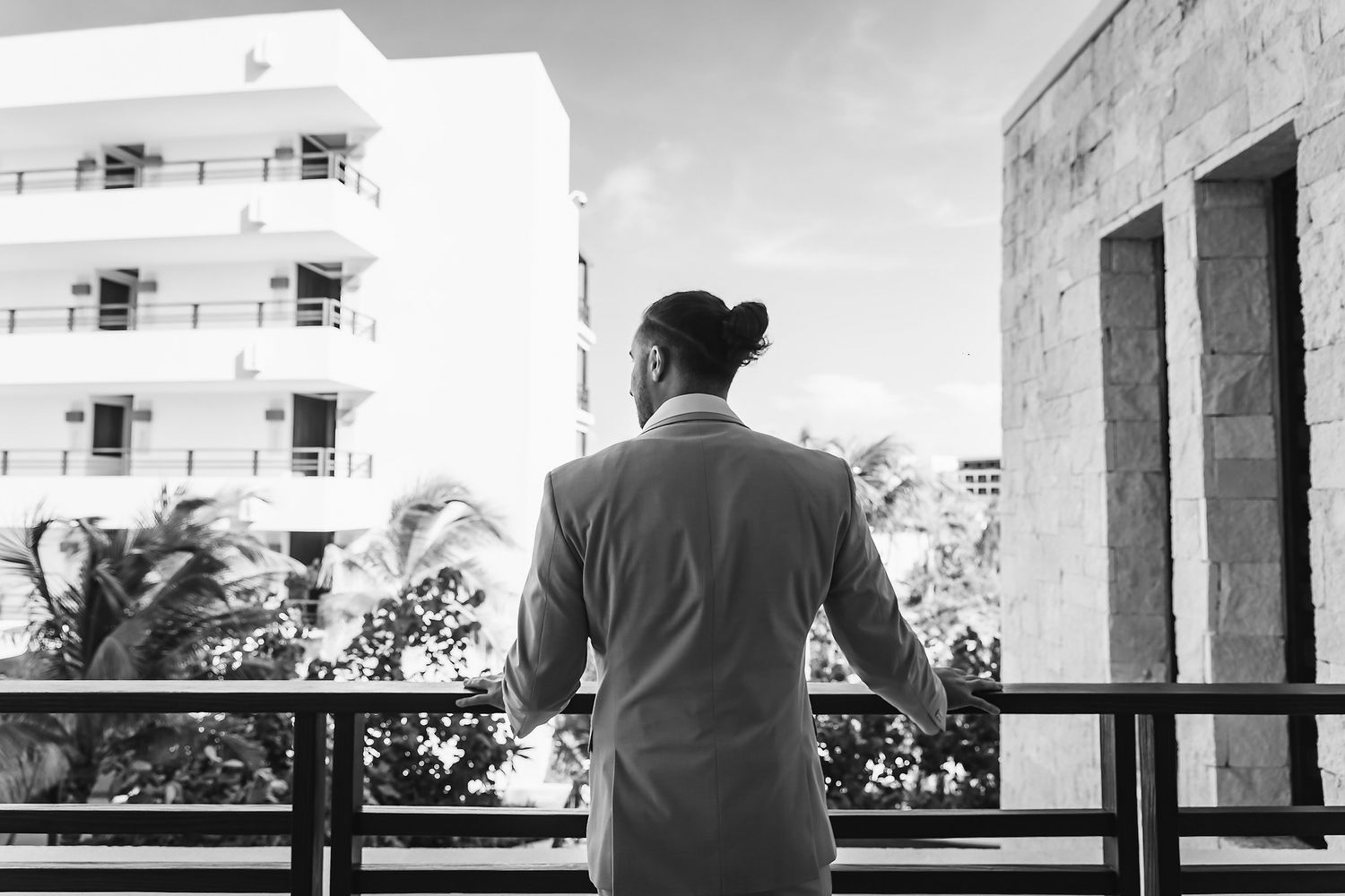 Man in suit standing on balcony overlooking tropical buildings, black and white photography.