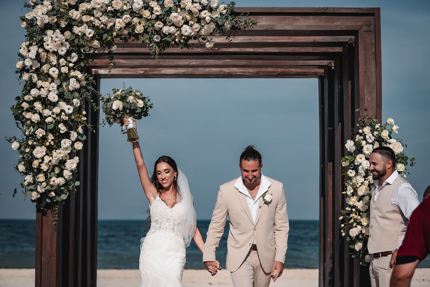 Bride and groom celebrate after beach wedding ceremony under floral arch with white roses, ocean backdrop.