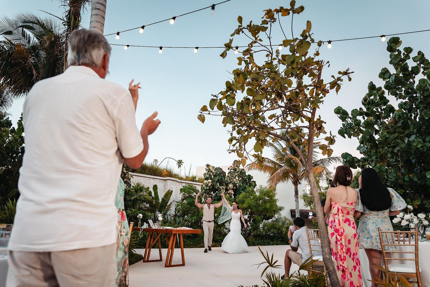 Guests celebrate an outdoor wedding reception with string lights and tropical trees at a beachside venue.