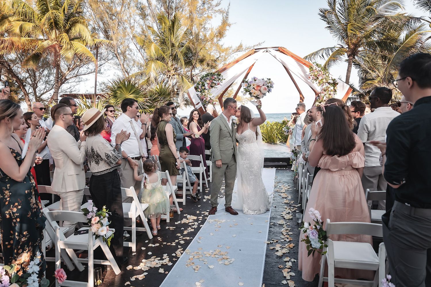 Newlyweds kiss at outdoor beach wedding ceremony while guests cheer under floral arch with palm trees in background.