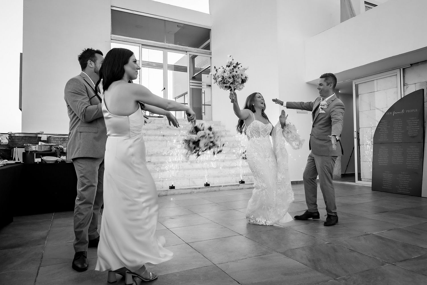 Black and white photo of a joyful wedding party celebrating indoors, with bride raising bouquet.