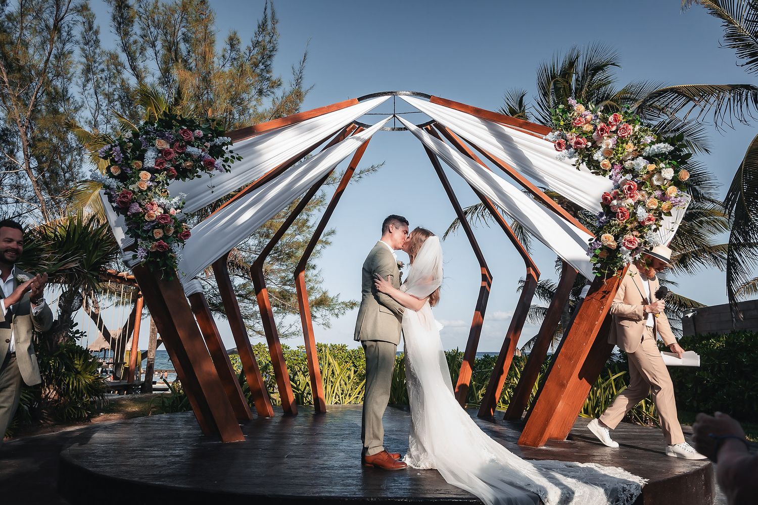 Couple kissing under geometric wooden arch with floral decor at a tropical beach wedding ceremony.