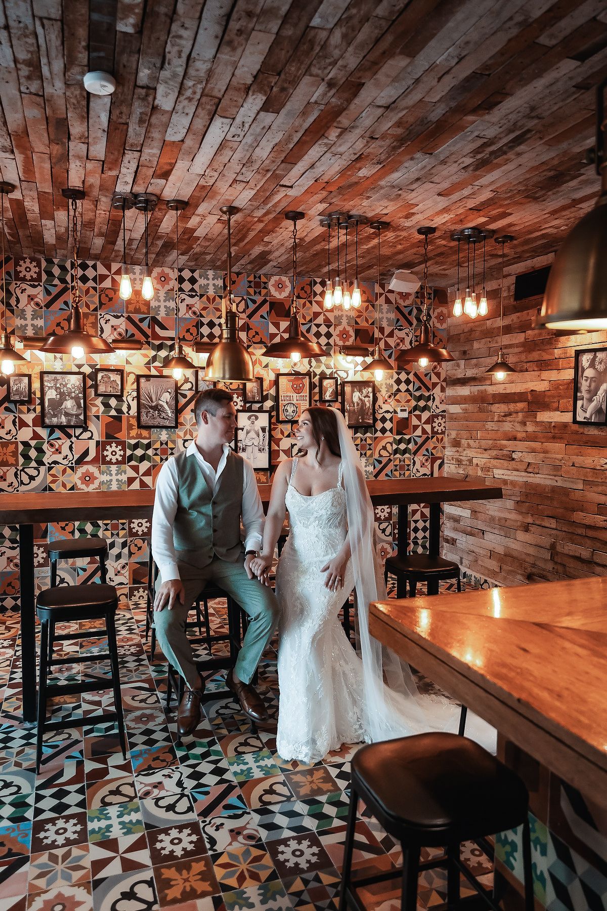 Wedding couple holding hands in a rustic bar with colorful mosaic tiles and warm Edison lights.