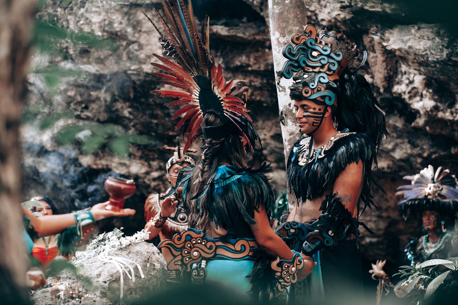 Group of performers in detailed Mayan ritual costumes and headdresses gathered near rocky surroundings.