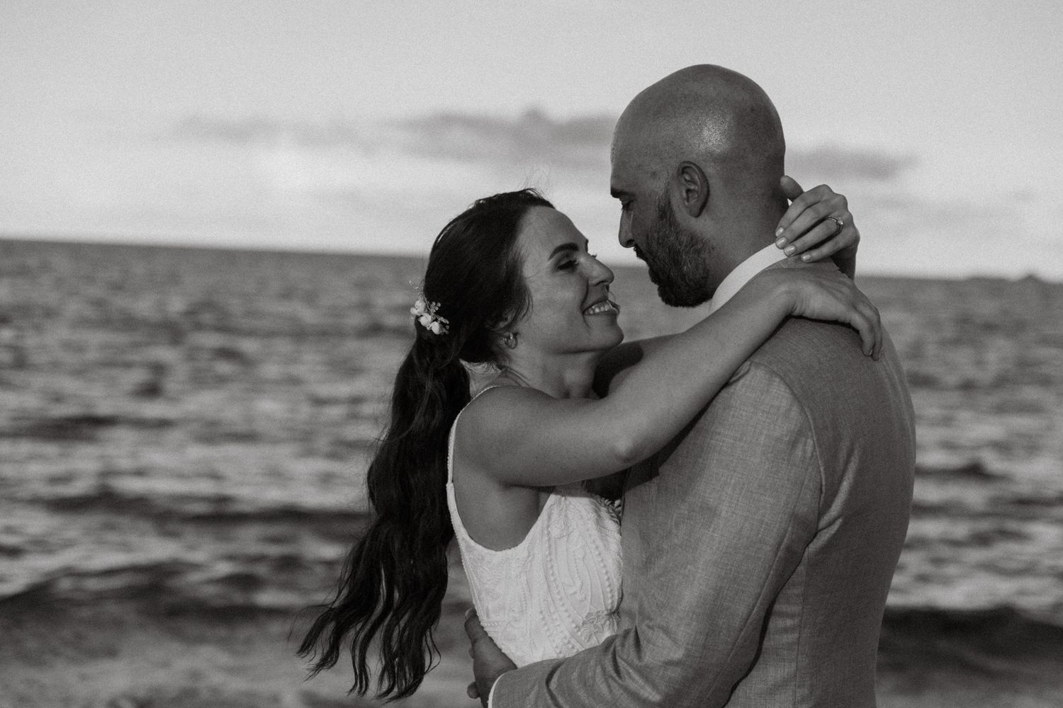 Black and white photo of a couple embracing and smiling at each other on a beach at sunset.