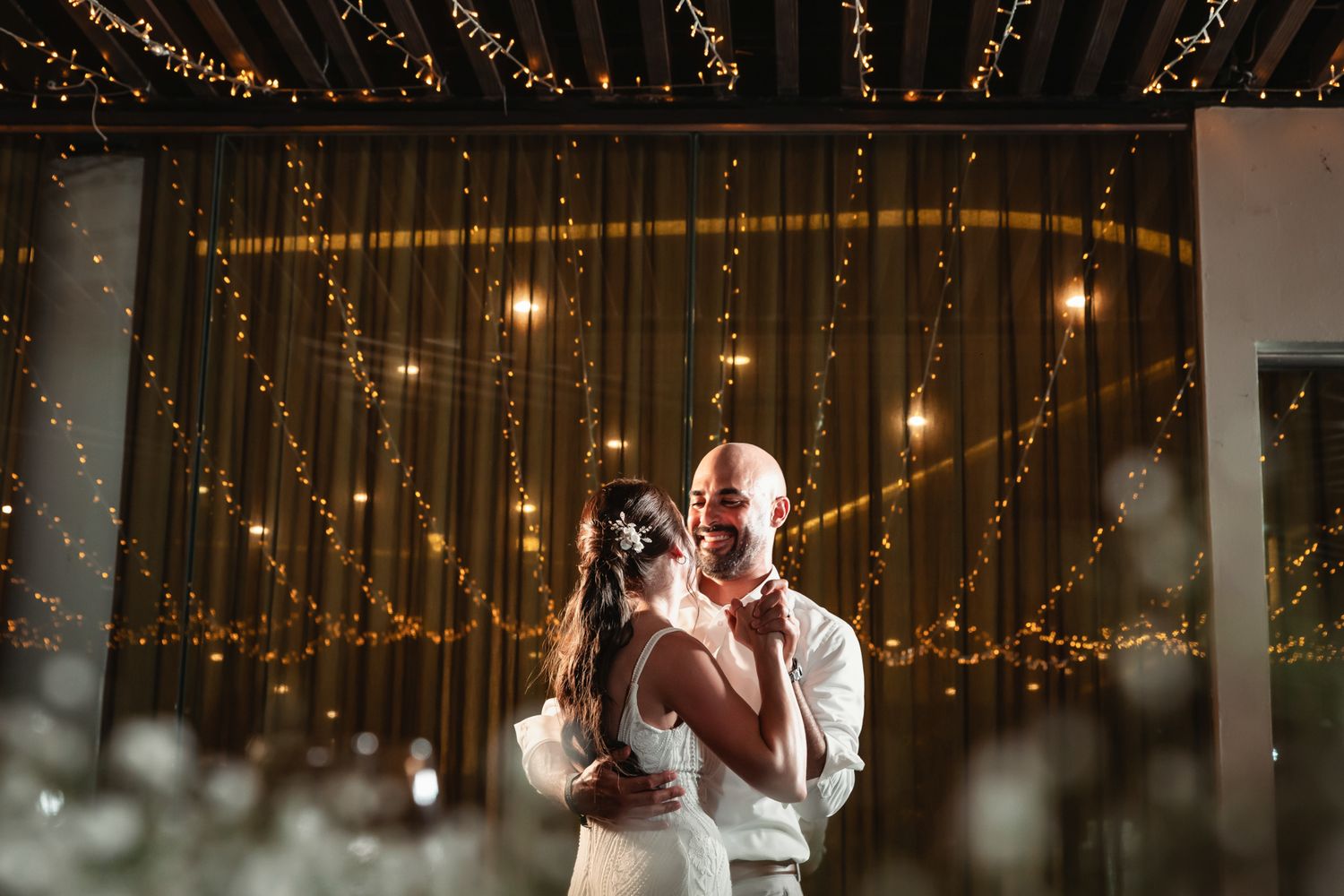 Couple sharing first dance under warm fairy lights and draped curtains at romantic wedding reception.