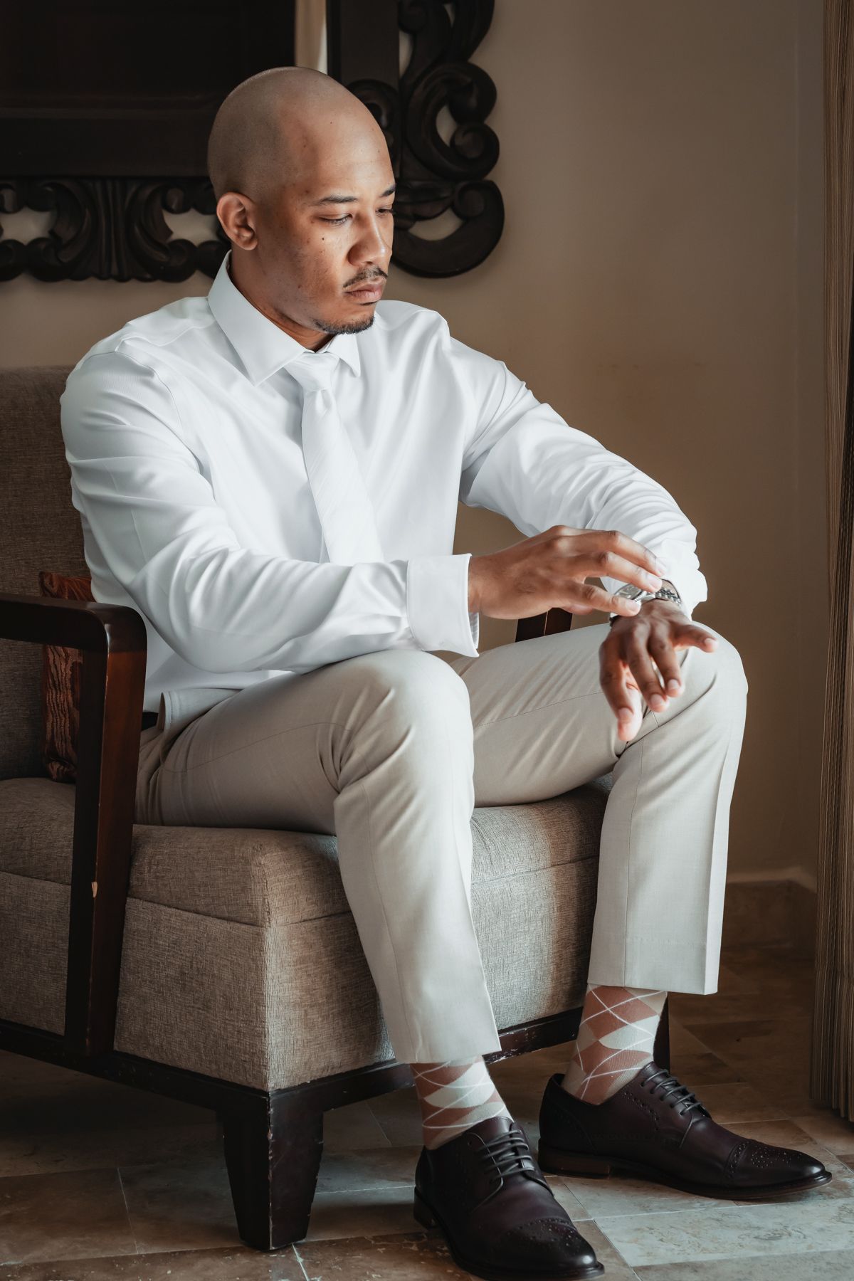Groom in white shirt and khaki pants sits in armchair adjusting cufflinks on wedding day.