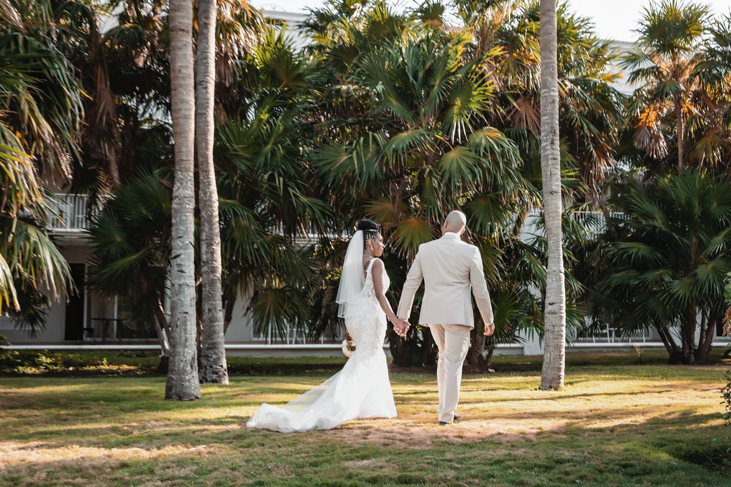 Bride and groom walk hand-in-hand through a sunlit tropical garden with tall palm trees at an outdoor wedding venue.