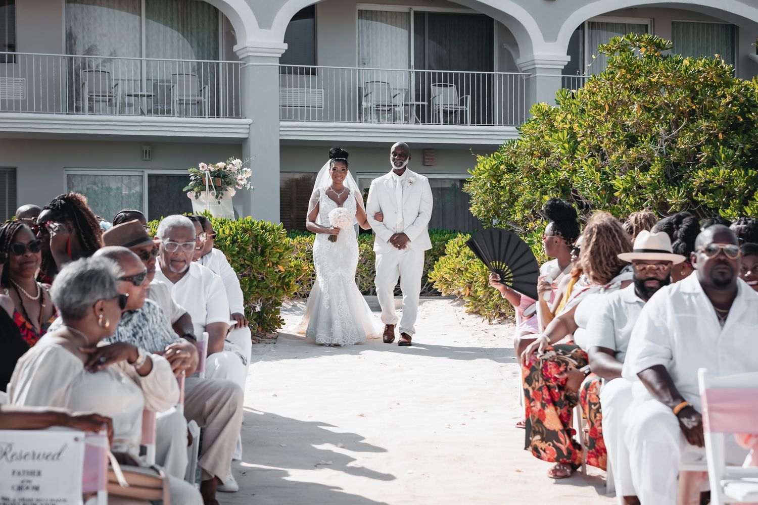 Outdoor wedding ceremony with bride and groom in white attire walking down aisle, guests seated, hotel backdrop.
