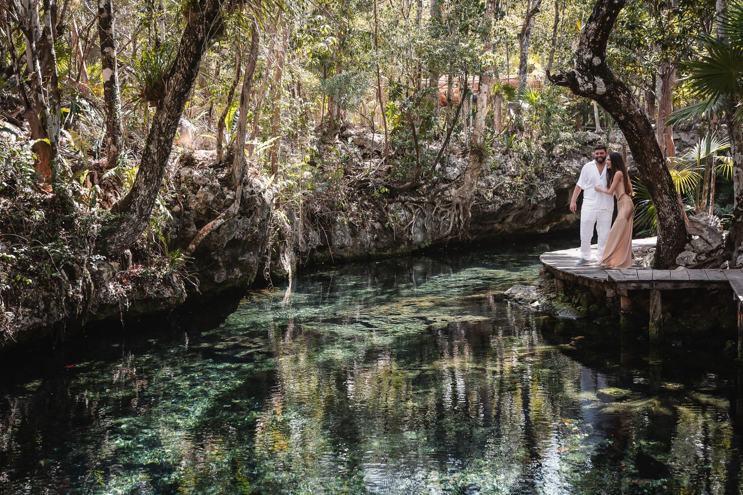 Couple in white attire standing on a wooden dock beside a serene cenote surrounded by lush jungle trees in Mexico.