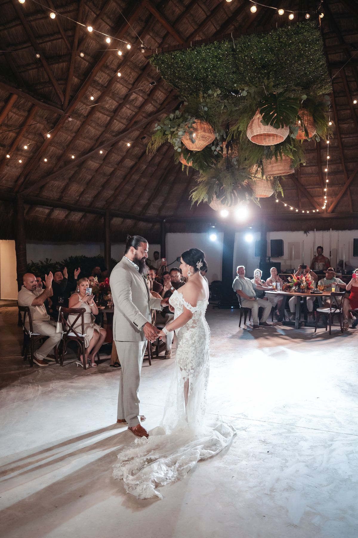 Bride and groom share first dance under tropical floral ceiling installation with string lights at outdoor wedding reception.