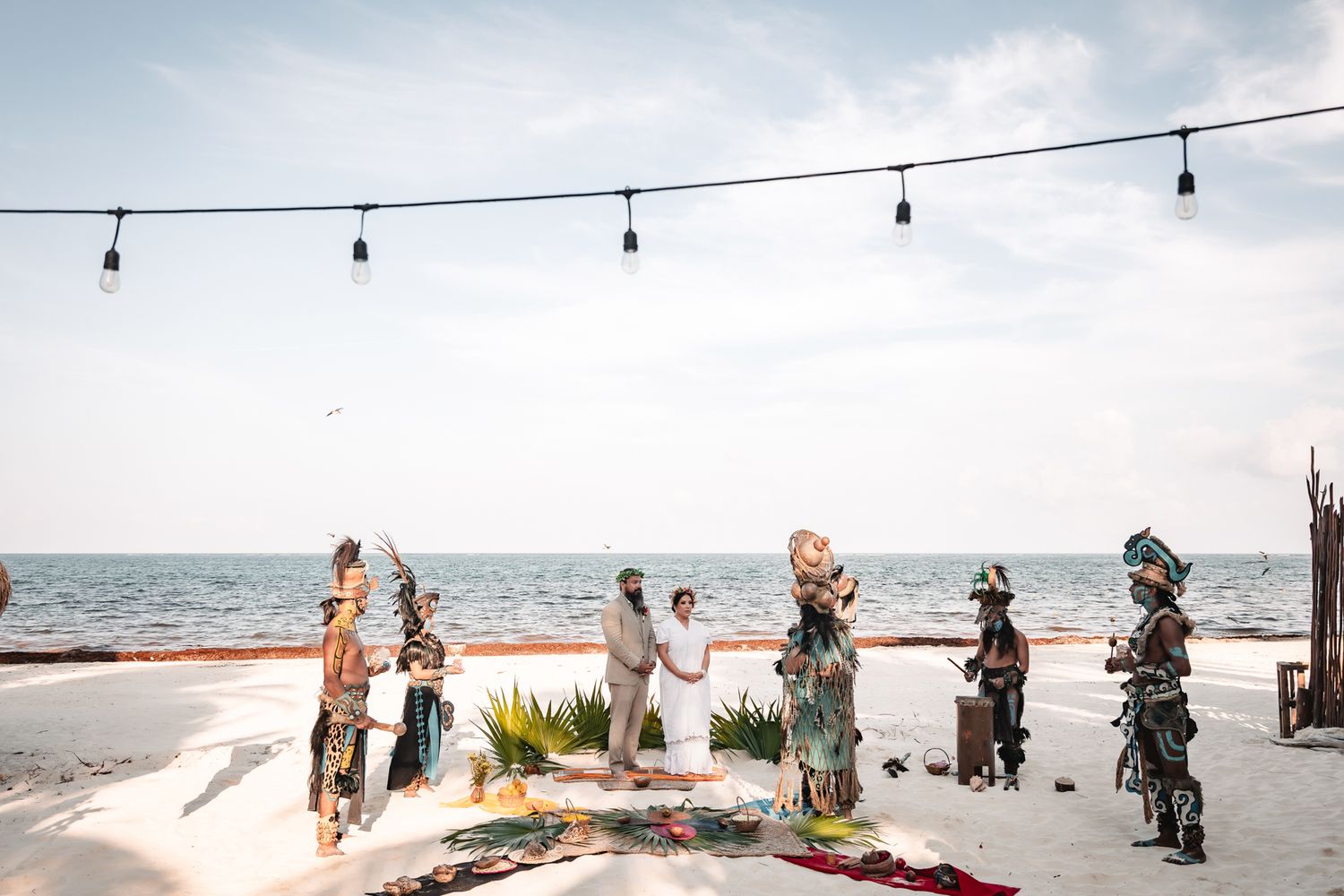 Bohemian beach wedding ceremony with colorful costumes, string lights, and ocean backdrop in a tropical destination.