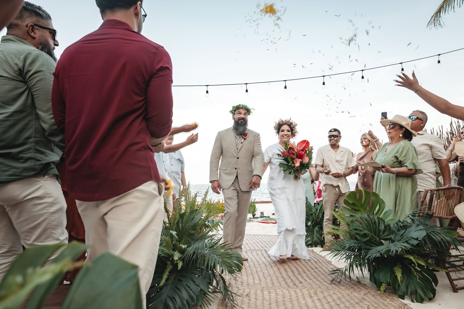 Newlywed couple walks down the aisle at an outdoor beach wedding ceremony with guests cheering and tossing confetti.