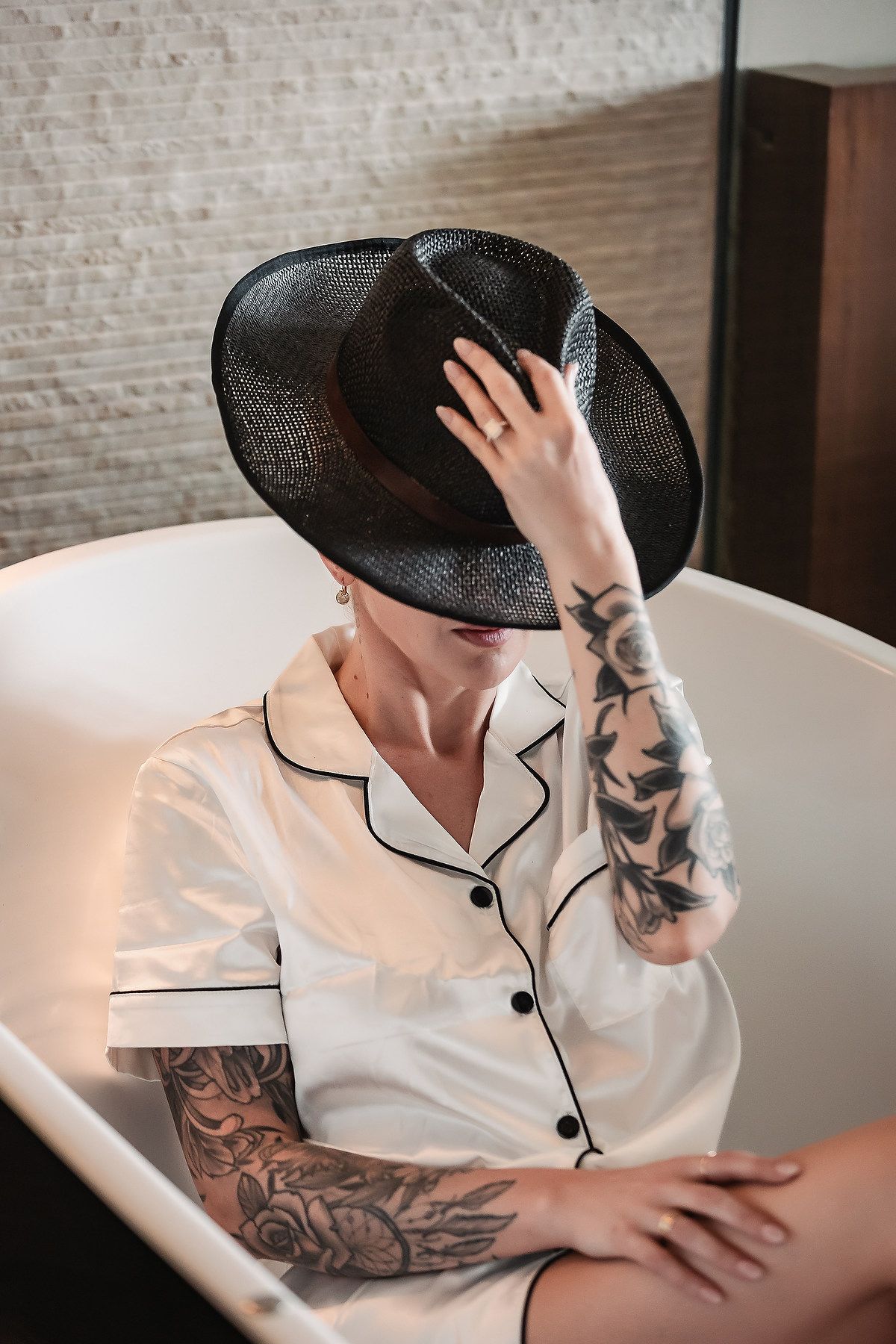 Person in white pajama top with black trim and wide-brimmed black hat poses in boudoir style setting.
