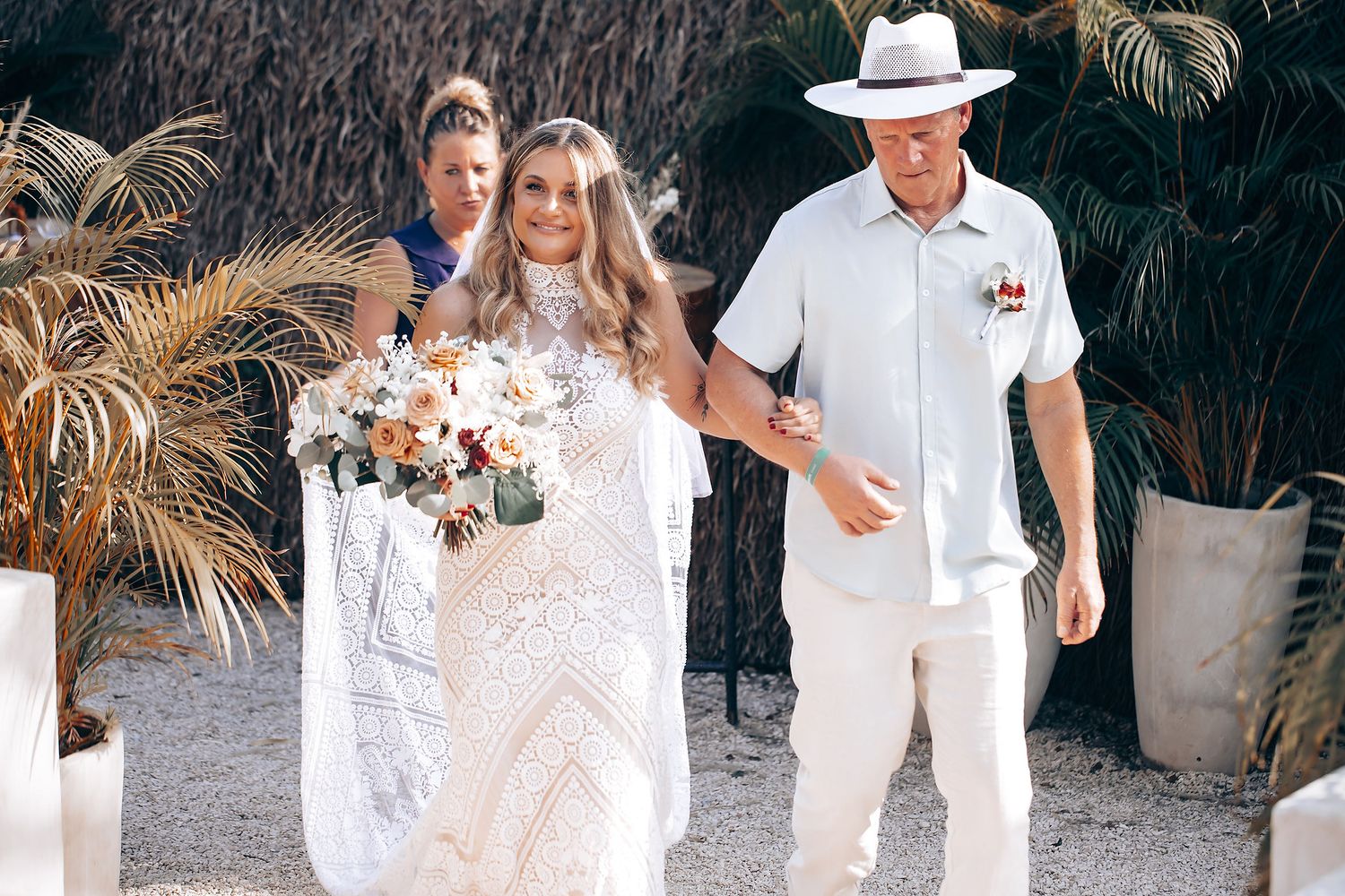 A tropical beach wedding celebration with guests in white and cream attire standing under palm trees.