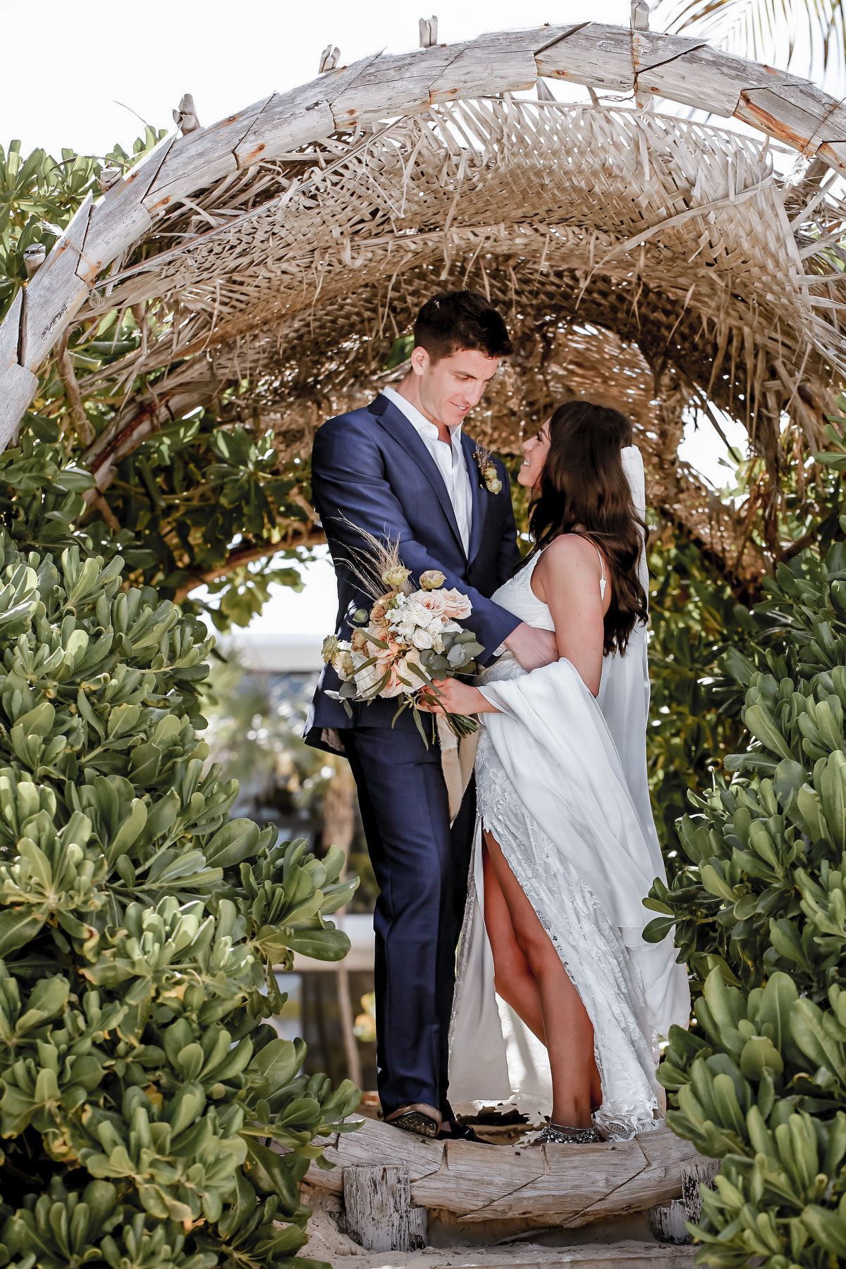Couple embraces under a rustic beach arbor surrounded by tropical foliage.