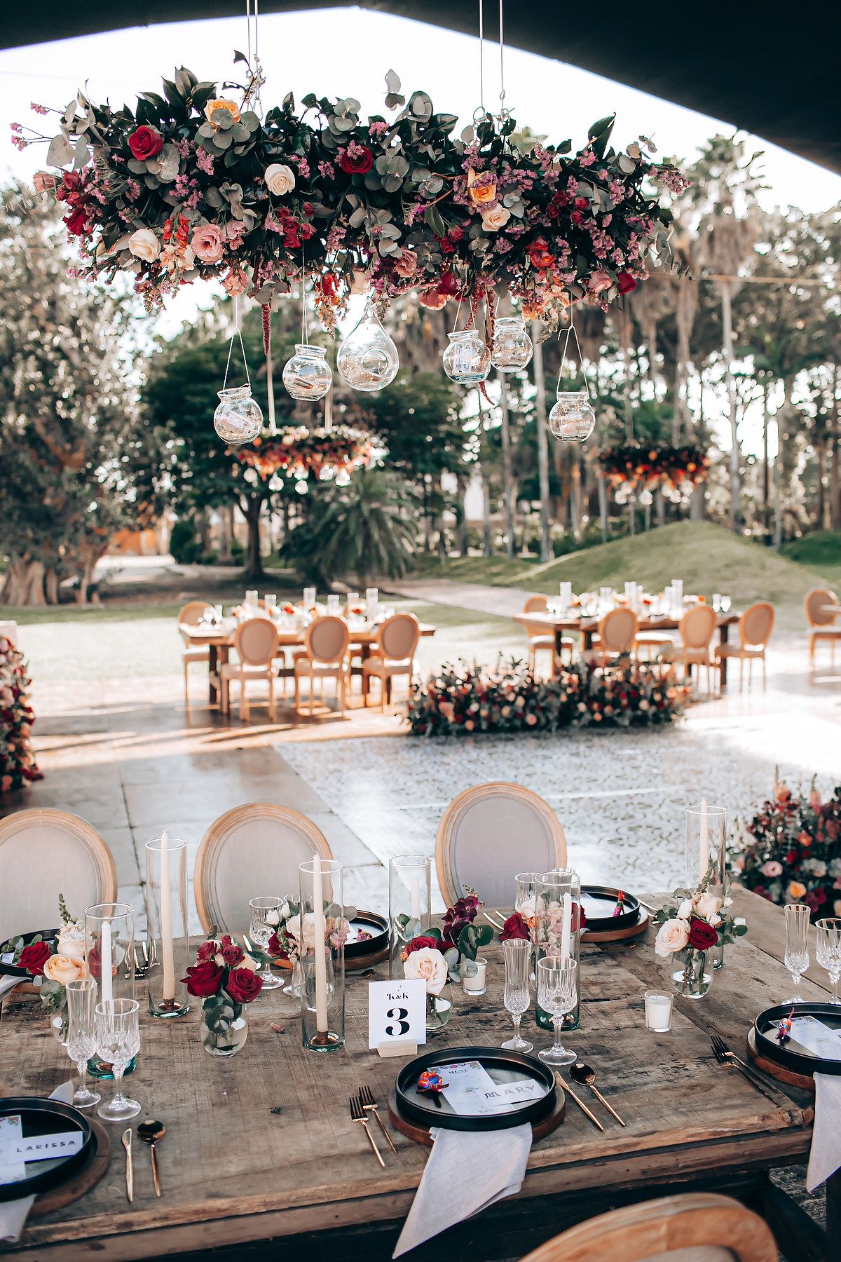 Rustic outdoor wedding reception table settings with wooden chairs and floral arrangements under hanging glass orbs at dusk.