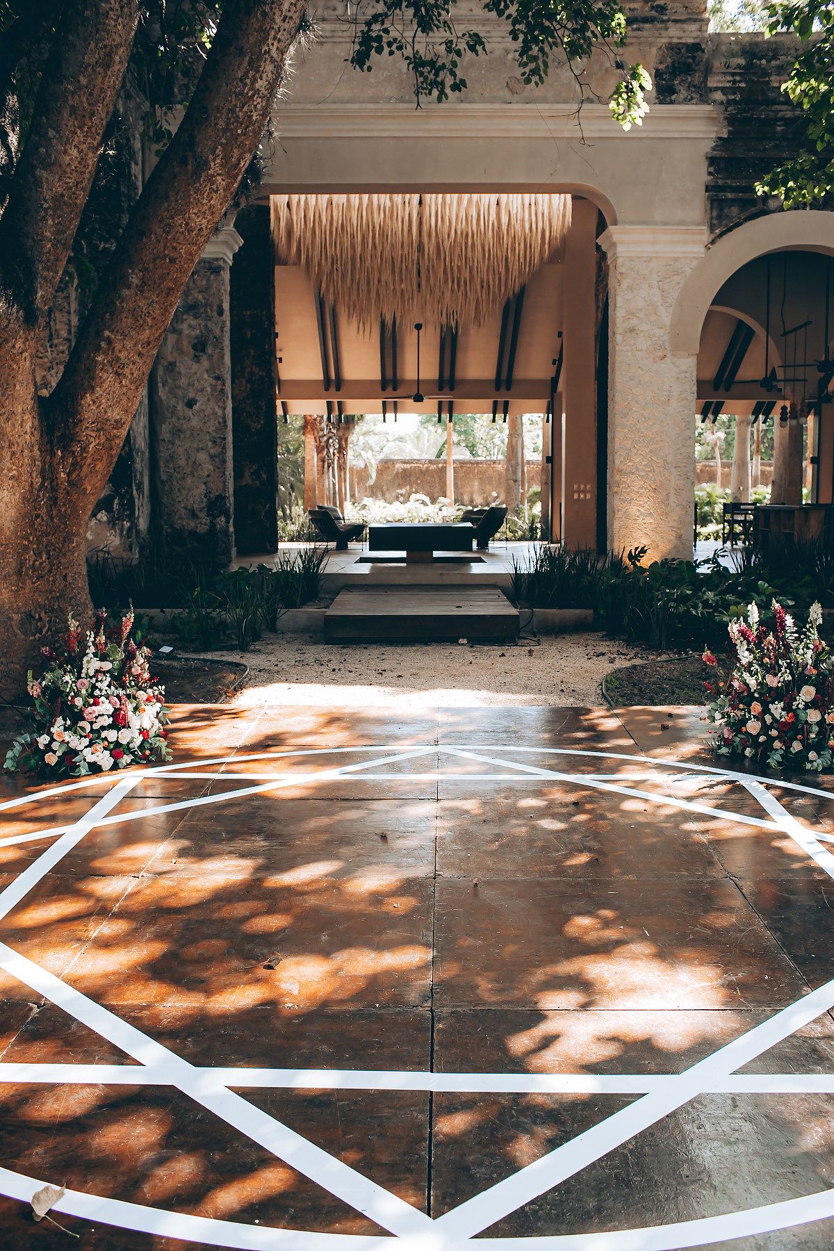 Elegant outdoor wedding venue with geometric floor pattern and rustic canopy framed by natural tree shadows.