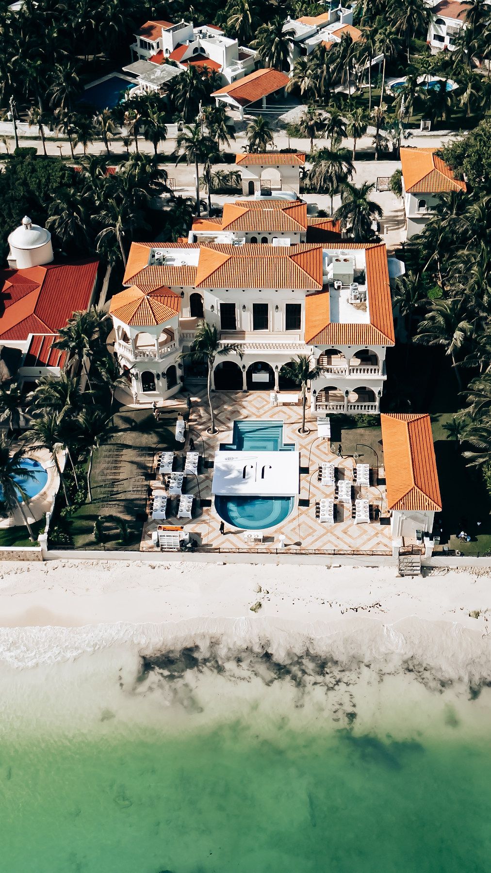 Aerial view of luxury beachfront homes with terracotta roofs and swimming pools along turquoise waters.