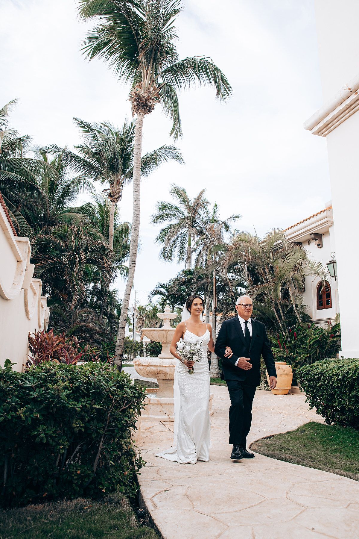 A wedding ceremony takes place on a tropical path lined with palm trees and white architecture.