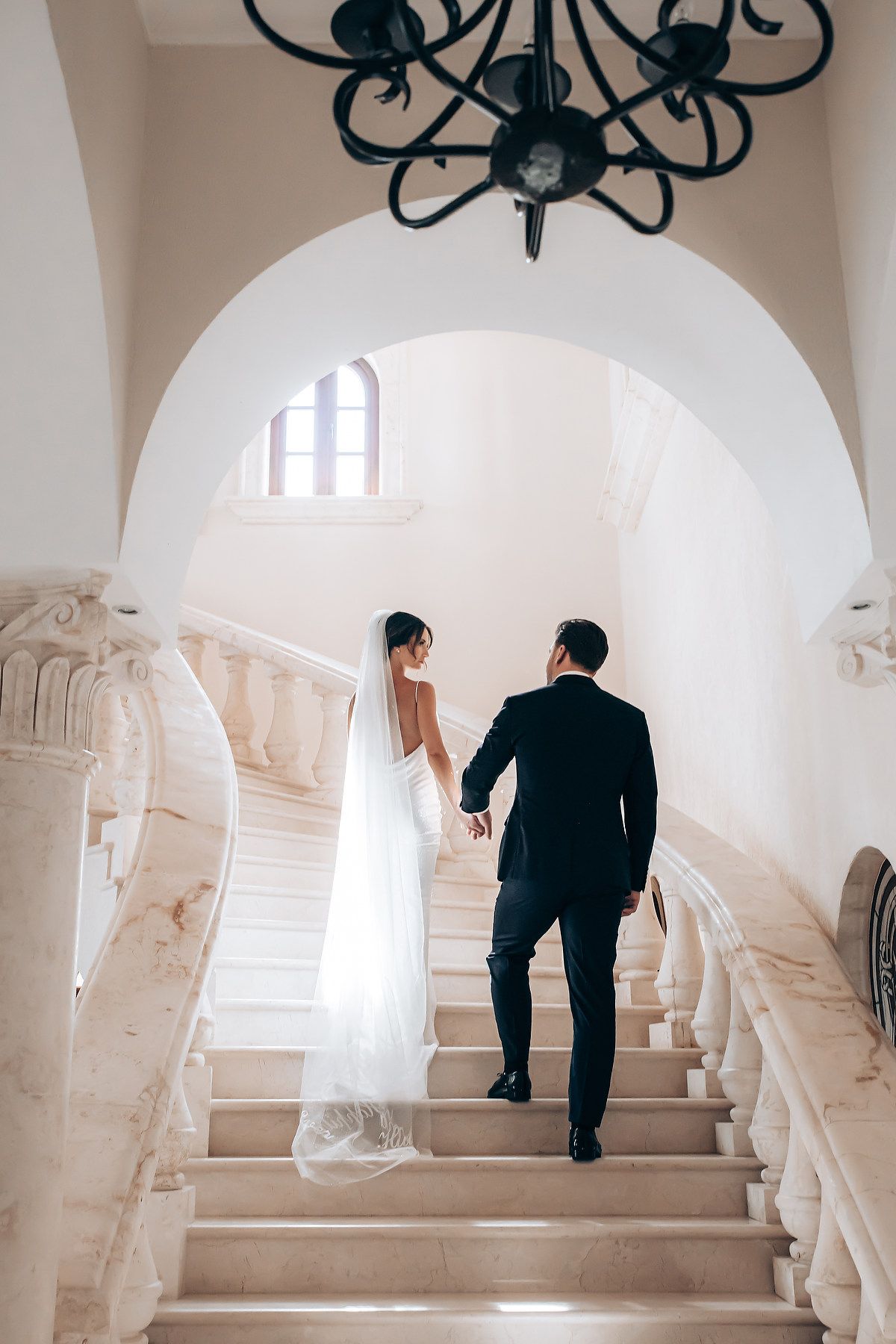 A couple walks up elegant white marble stairs with ornate railings and arched architecture in a romantic setting.