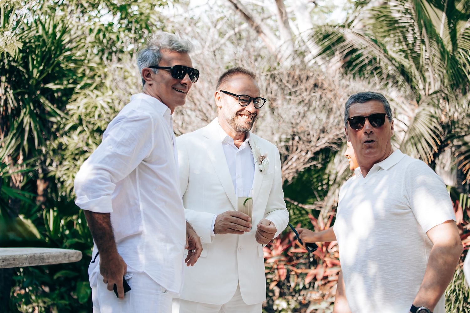 Three people wearing white outfits and sunglasses stand together outdoors with tropical foliage in the background.