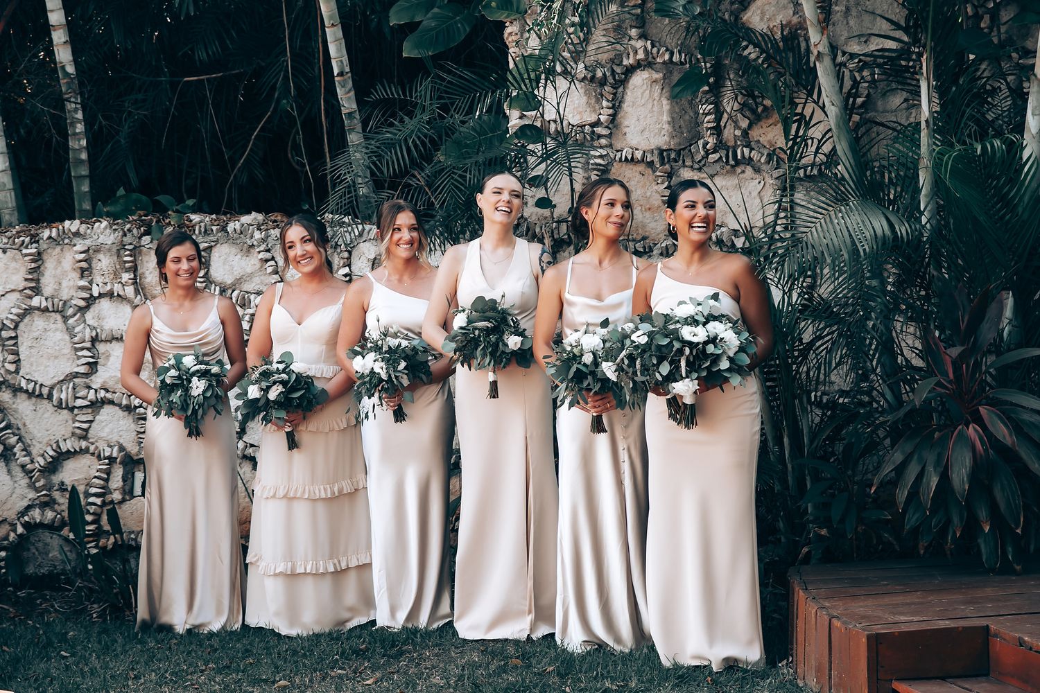 Bridesmaids in elegant cream-colored dresses pose together at an outdoor wedding venue.