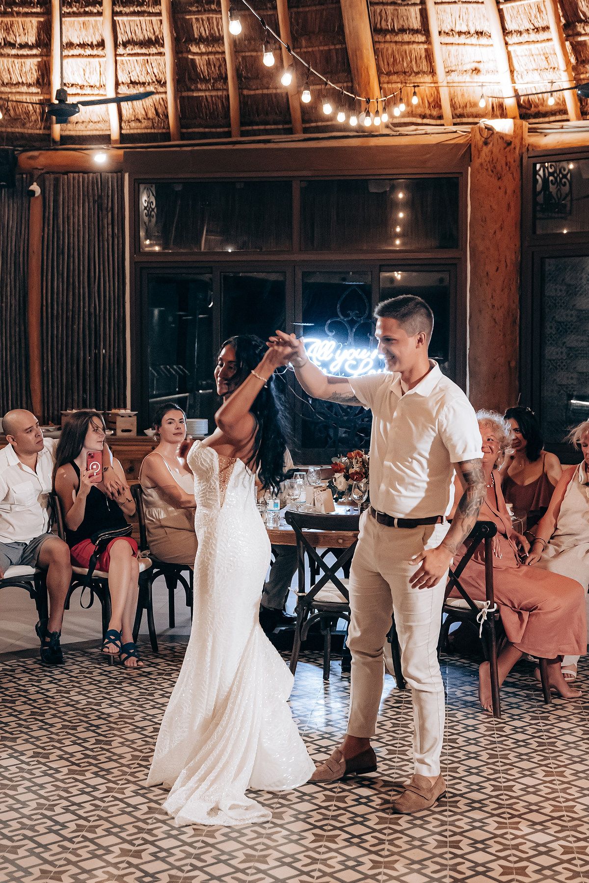 Wedding guests dancing on a patterned floor under a thatched roof palapa with string lights.