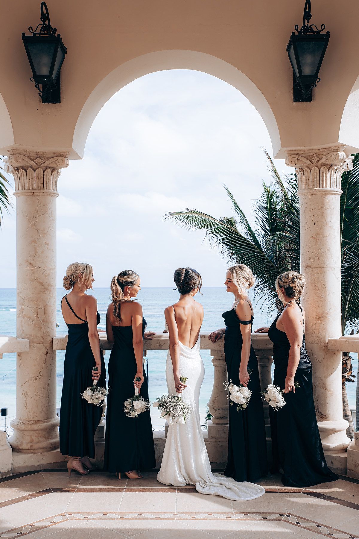 Bridesmaids in black dresses stand with bride under arched colonnade overlooking ocean.