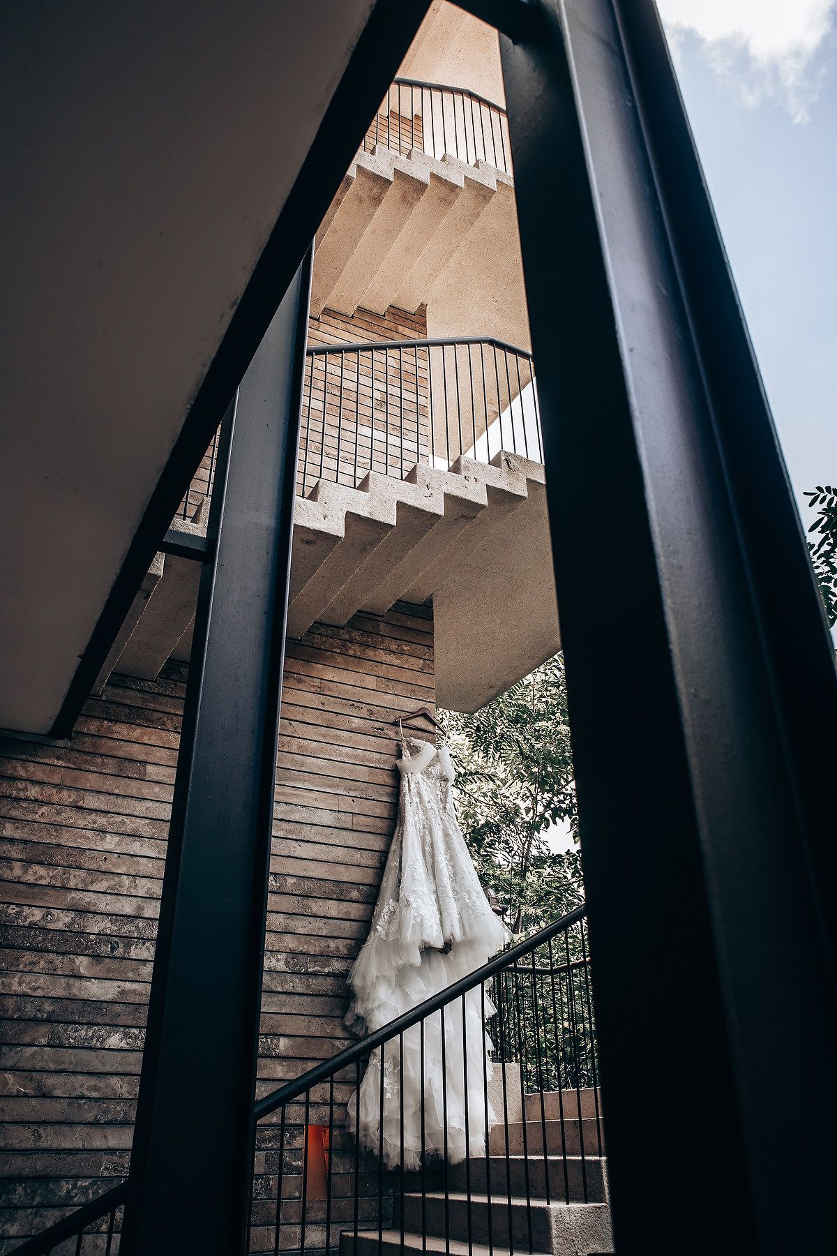 A white wedding dress hangs elegantly on a spiral staircase with dramatic architectural framing.